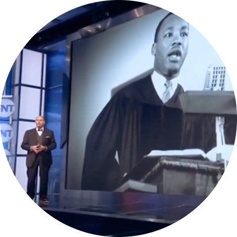 A tv host stands next to a large video of Martin Luther King Jr.