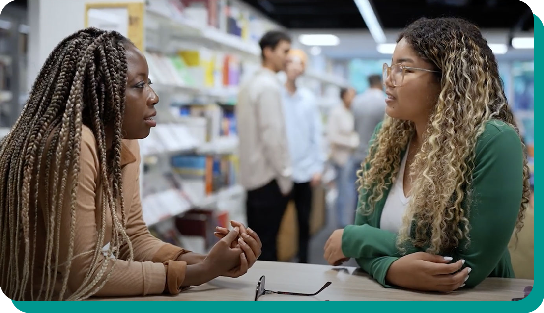 An African American woman with long braids speaking to a young woman with glasses and long wavy hair in a bookstore or library