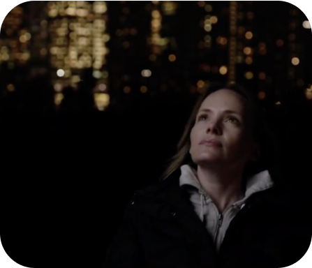 a woman wearing a warm hooded coat standing outside at night with lights from highrise buildings in the background