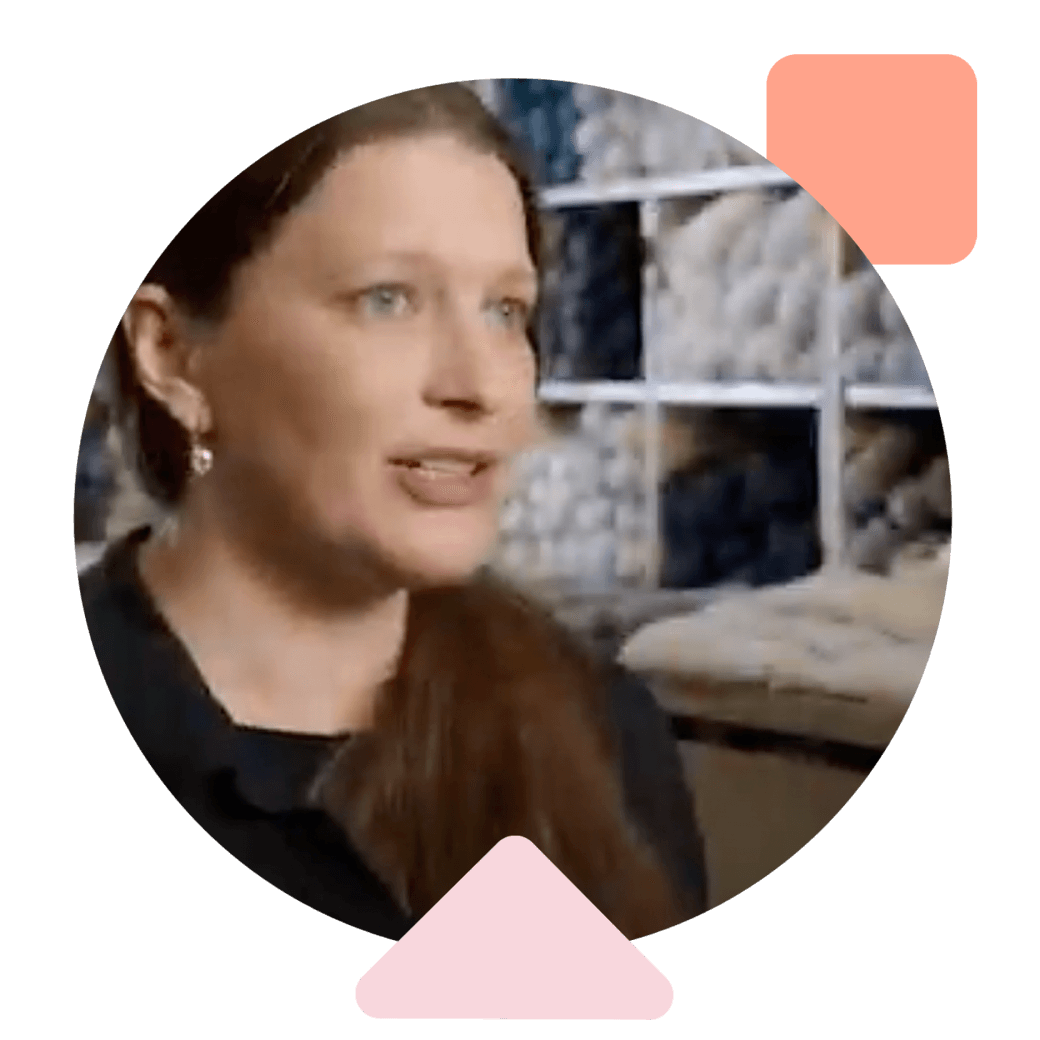 a woman with blue eyes and a side ponytail speaks in front of a shelf full of blue and white skeins of yarn